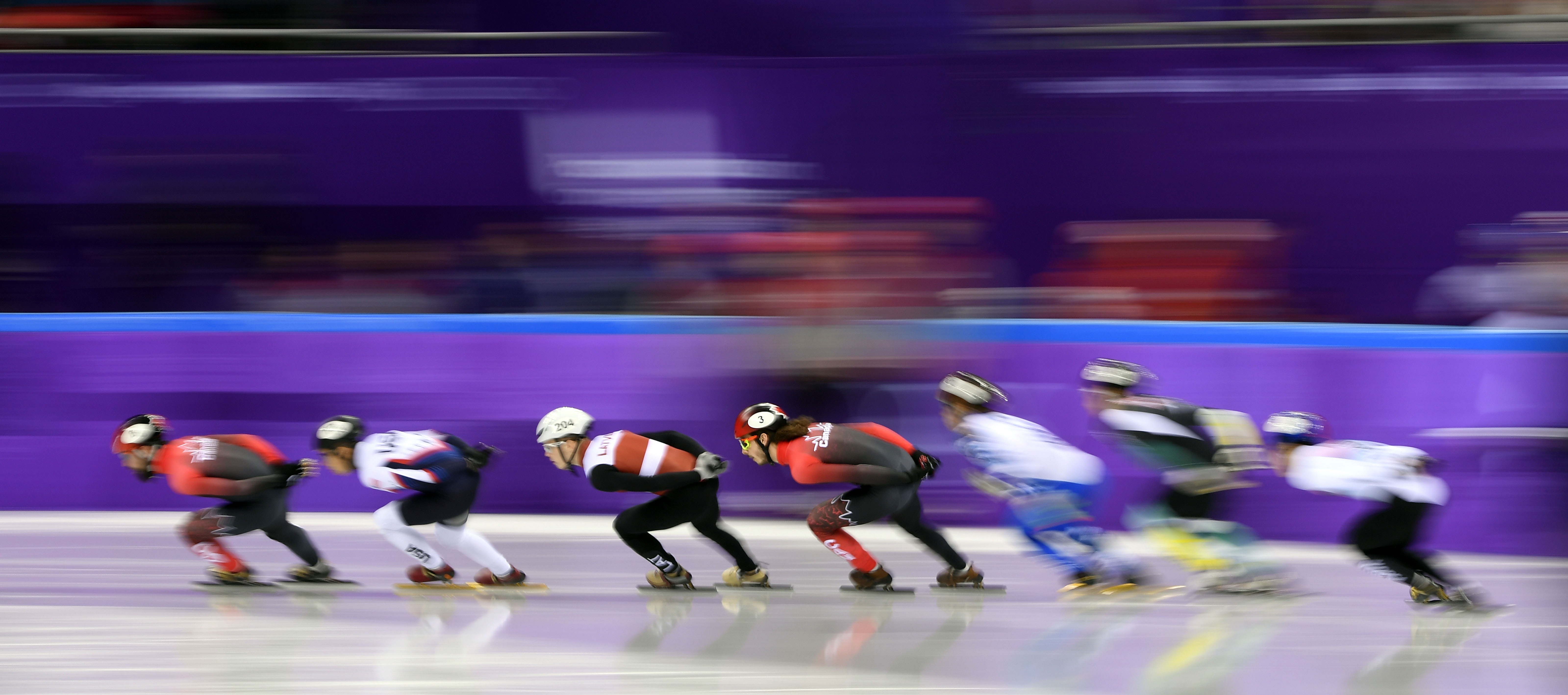 Plusieurs patineurs de vitesse en pleine course sur une patinoire, penchés vers l'avant pour prendre de la vitesse.